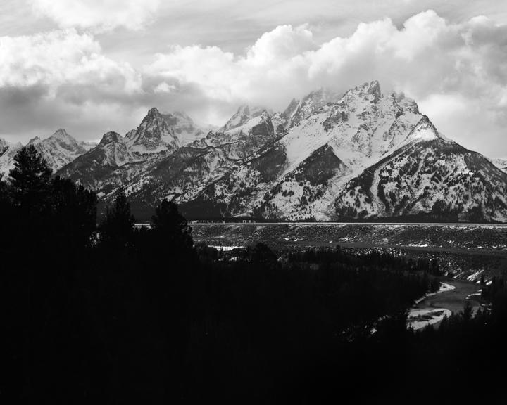 A black and white landscape shows snow-capped mountains under a cloudy sky, with a river and forest in the foreground.