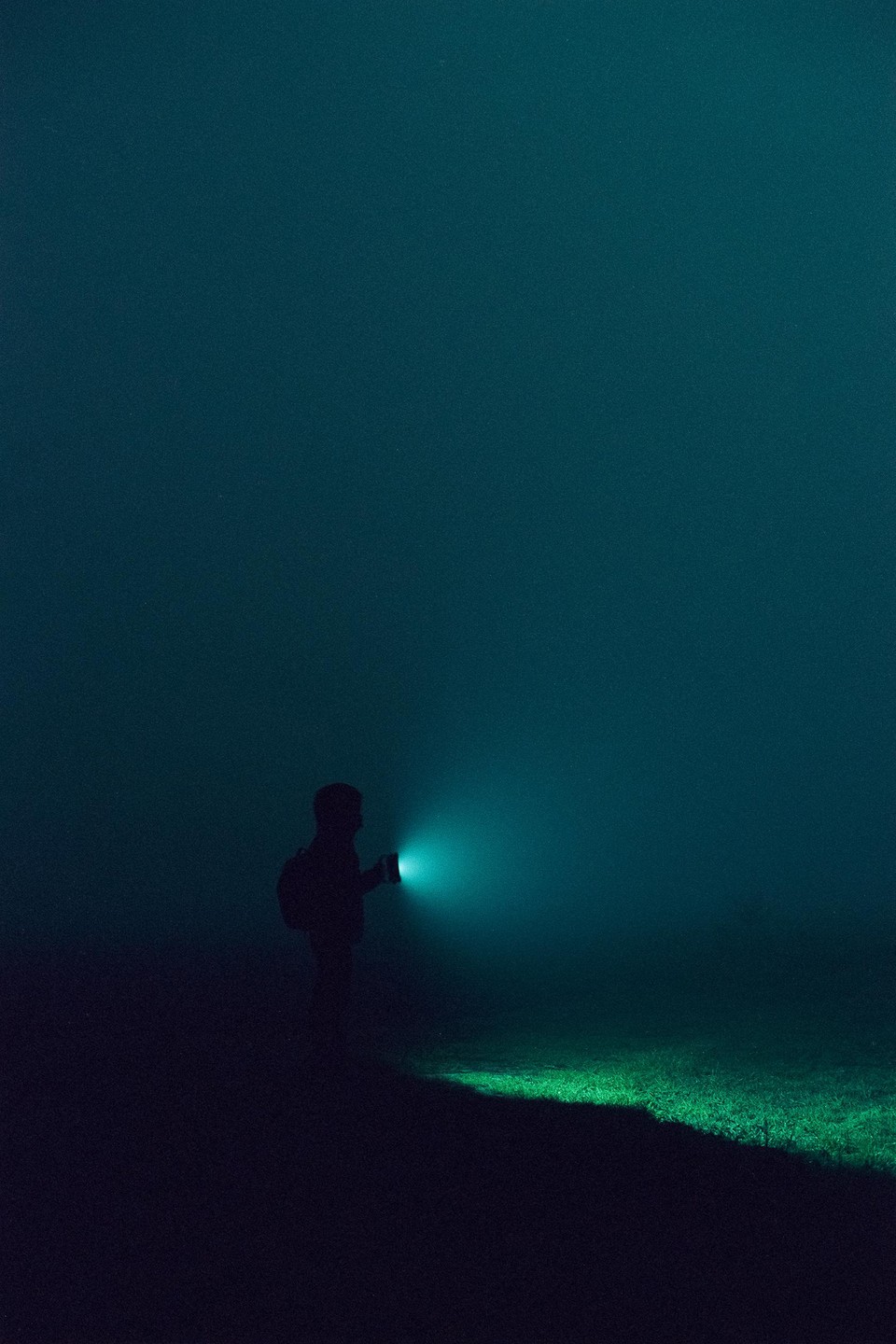 A man with a backpack holds a flashlight that illuminates the ground in front of him in a dark, foggy landscape.