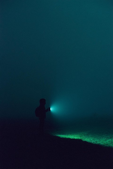 A man with a backpack holds a flashlight that illuminates the ground in front of him in a dark, foggy landscape.