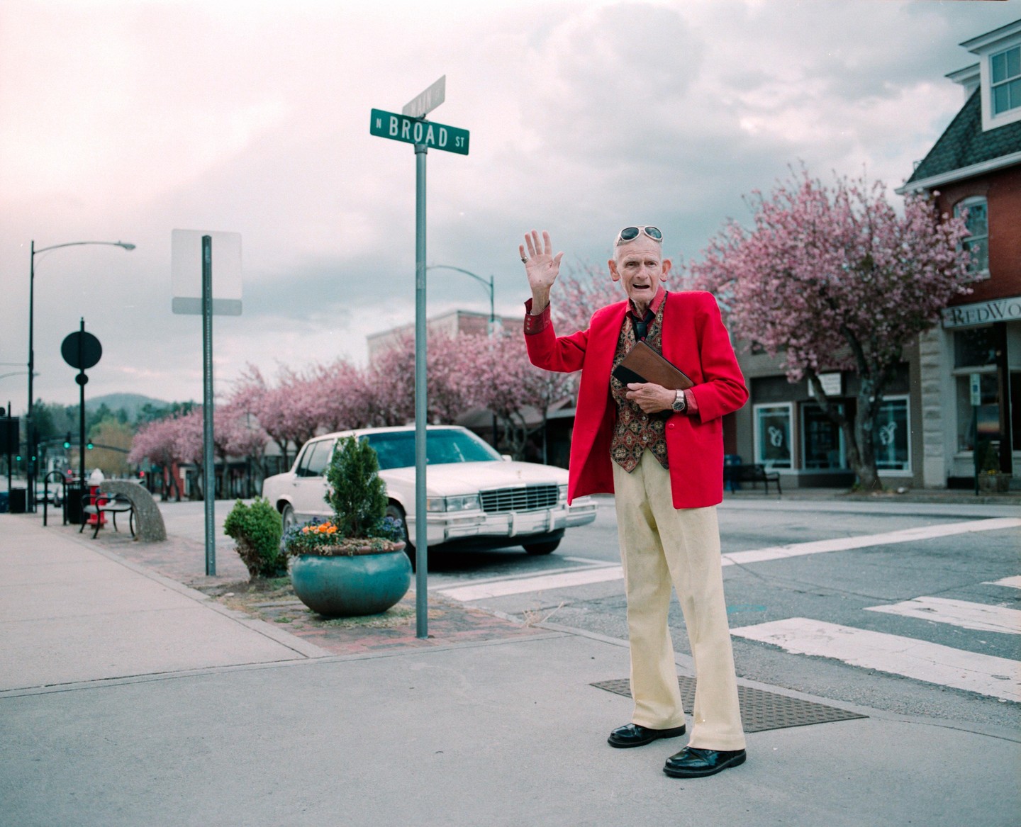 An elderly man in a red jacket waves from a street corner with blooming trees and a white car in the background.