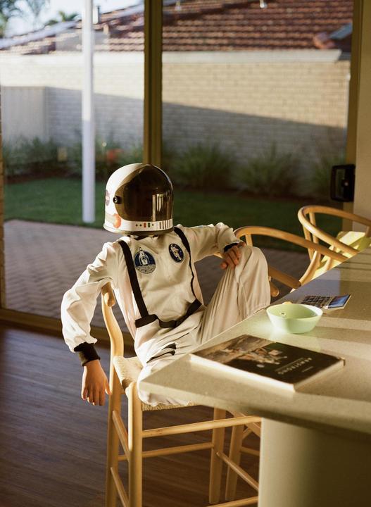 A child in an astronaut costume and helmet sits at a table with a book and a bowl, looking out a glass door to a yard.