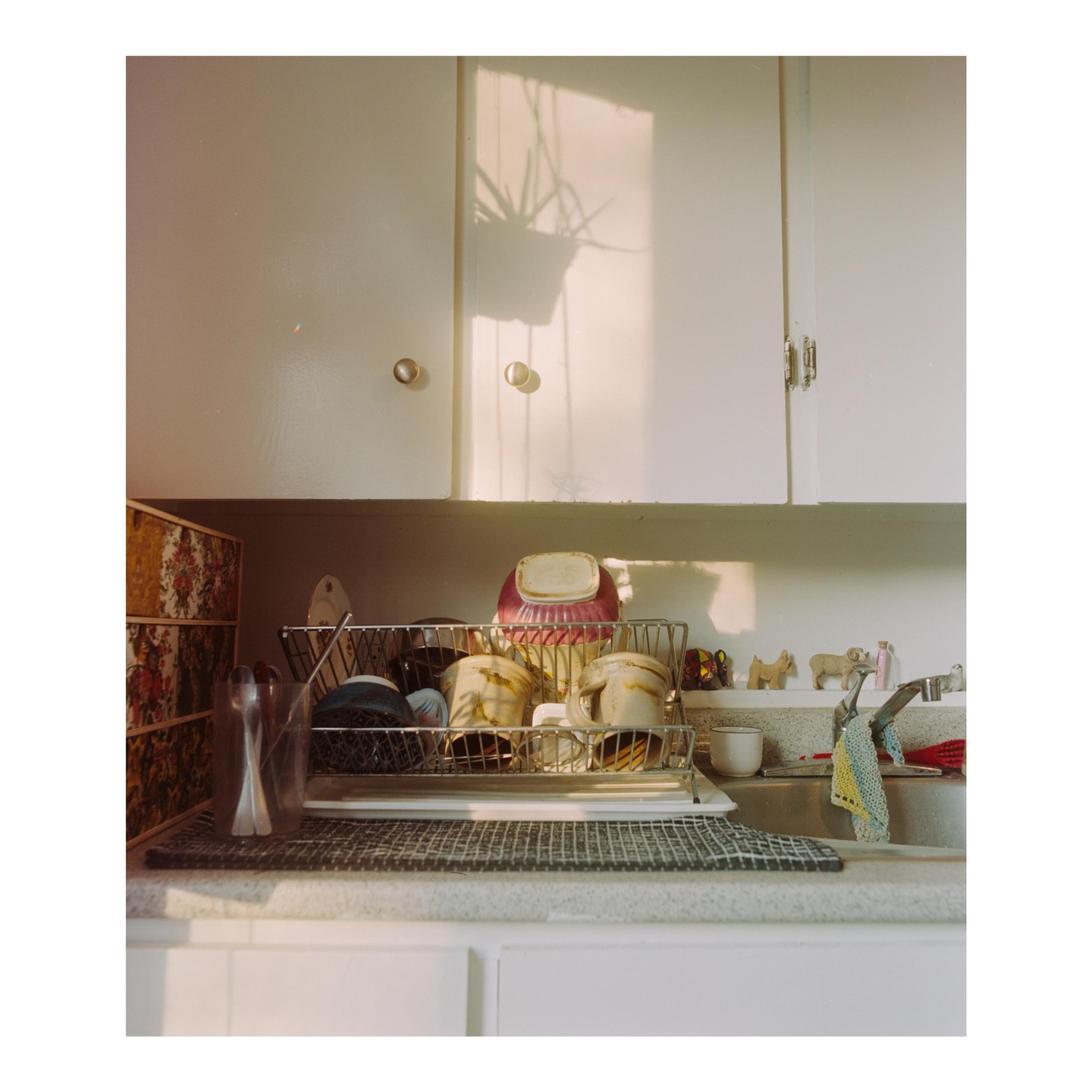 Sunlight casts shadows on white kitchen cabinets above a sink filled with dishes and decorative items.