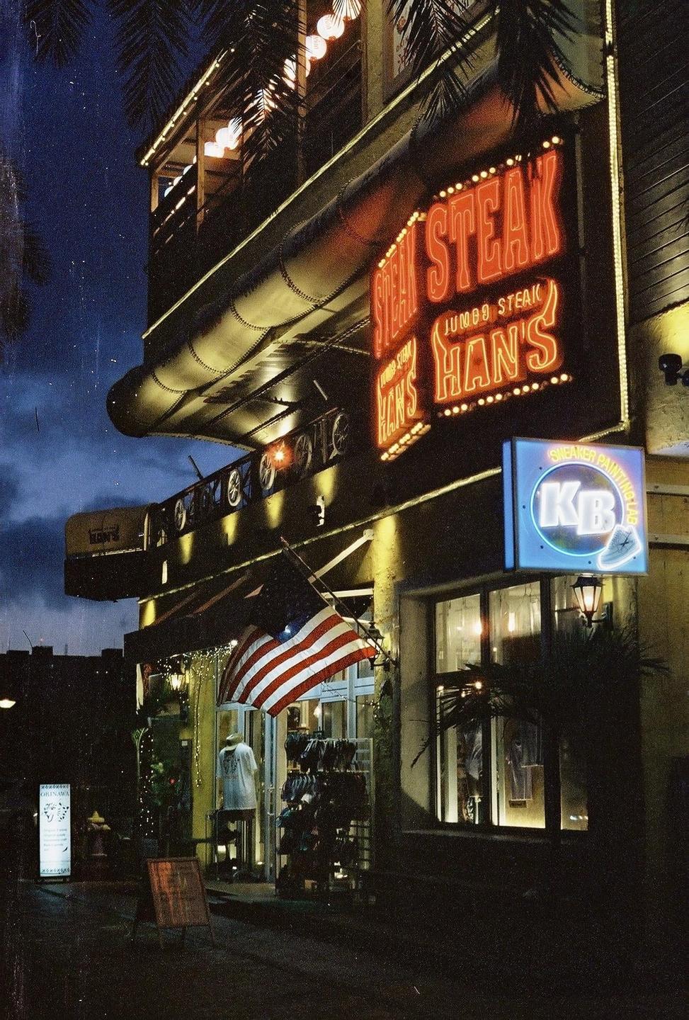A building with neon signs for "Steak Han's" and "KB" is illuminated at night, with an American flag hanging near the entrance.