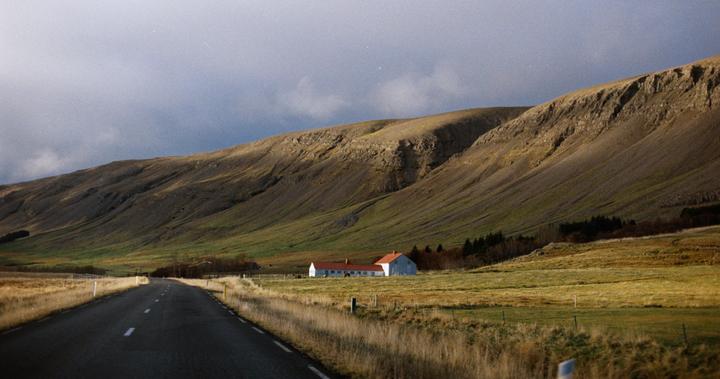 A road leads toward a white building with a red roof nestled at the base of a large, grassy mountain under a cloudy sky.