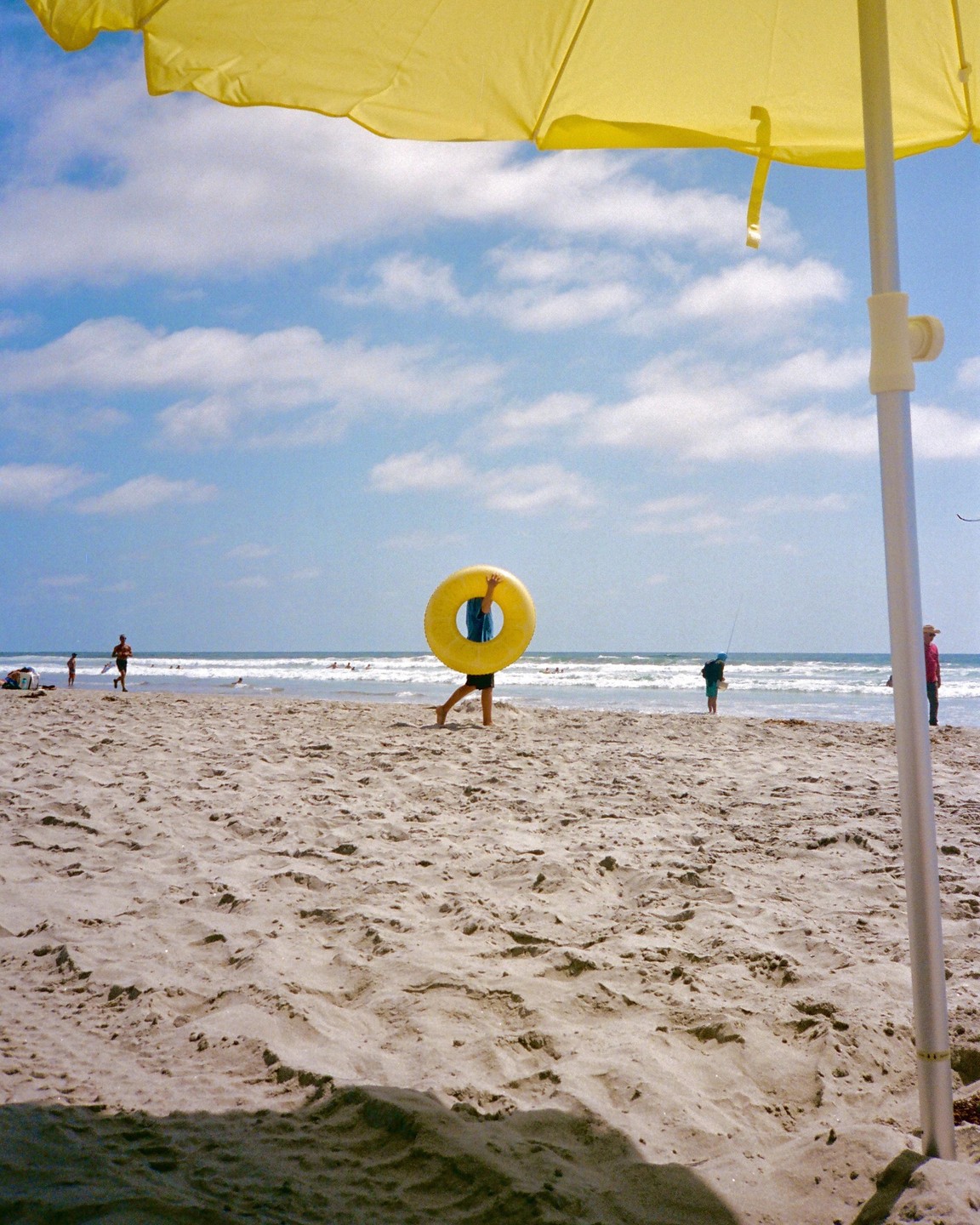 A person carries a yellow inflatable ring on a sandy beach under a yellow umbrella.