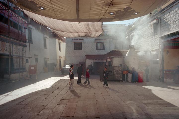 A group of people walk on a stone street with buildings and smoke in the background.