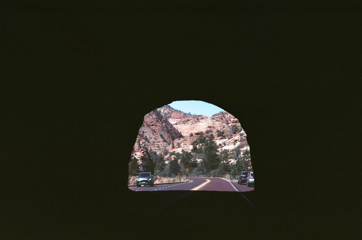 A view of a winding road through a mountainous landscape with cars, framed by the dark opening of a tunnel.