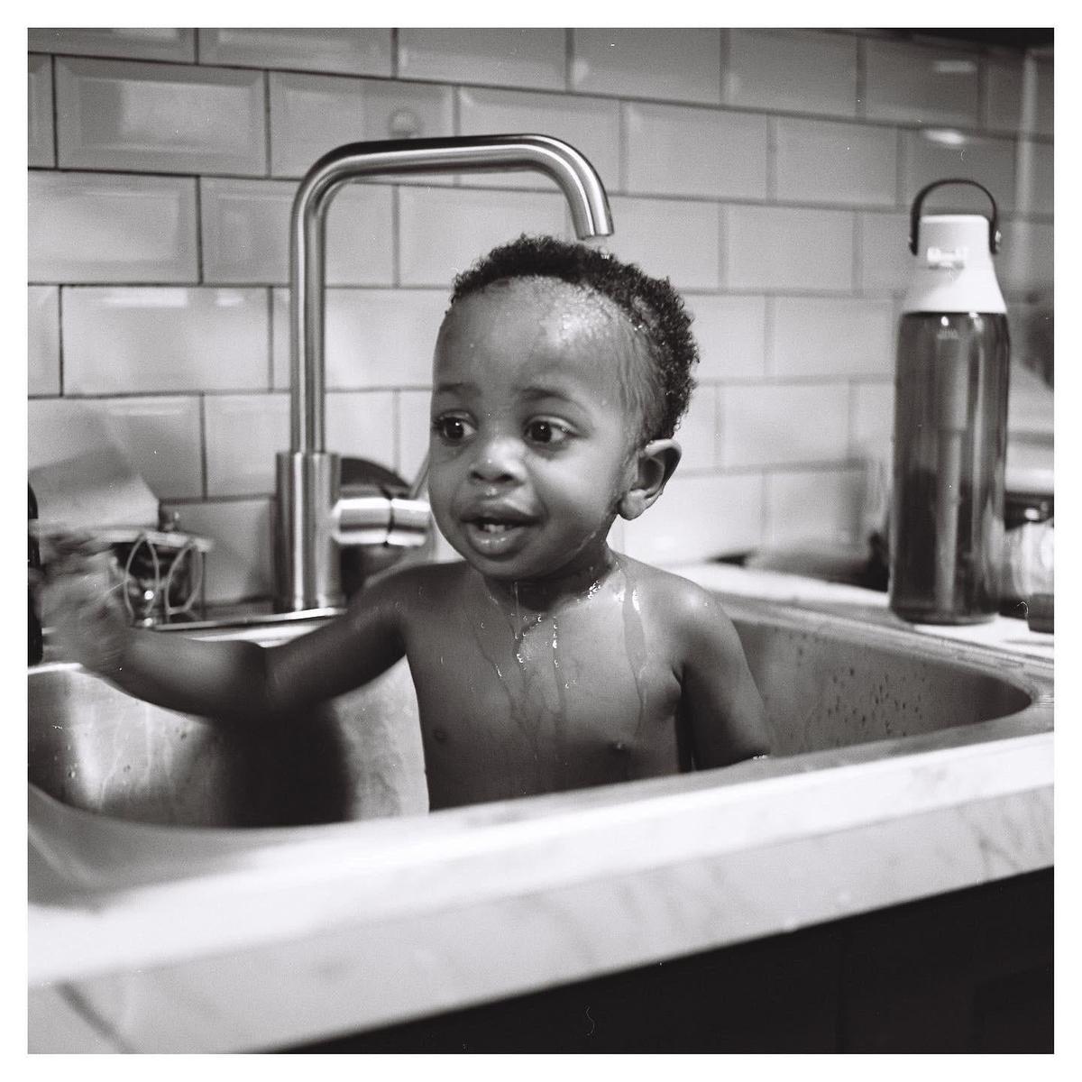 A child is standing in a kitchen sink with water dripping from their hair and chest.