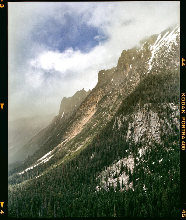 A mountain range covered in pine trees and patches of snow is partially obscured by clouds.