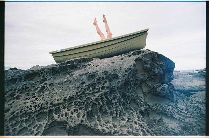 A small boat rests on a large, textured rock formation with a person's legs sticking out of the boat.