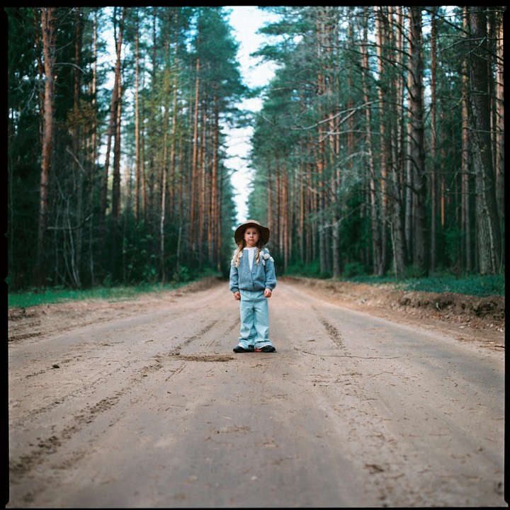 A young girl wearing a hat stands in the middle of a dirt road lined with tall trees.