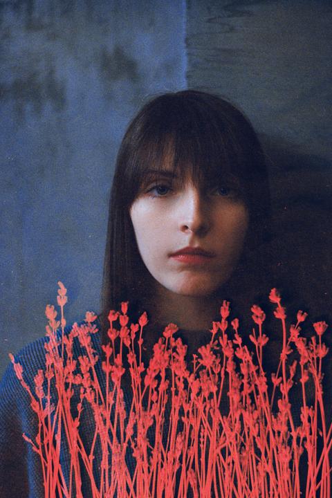 A woman with dark hair and bangs looks directly at the viewer, with bright red plant stems in the foreground.