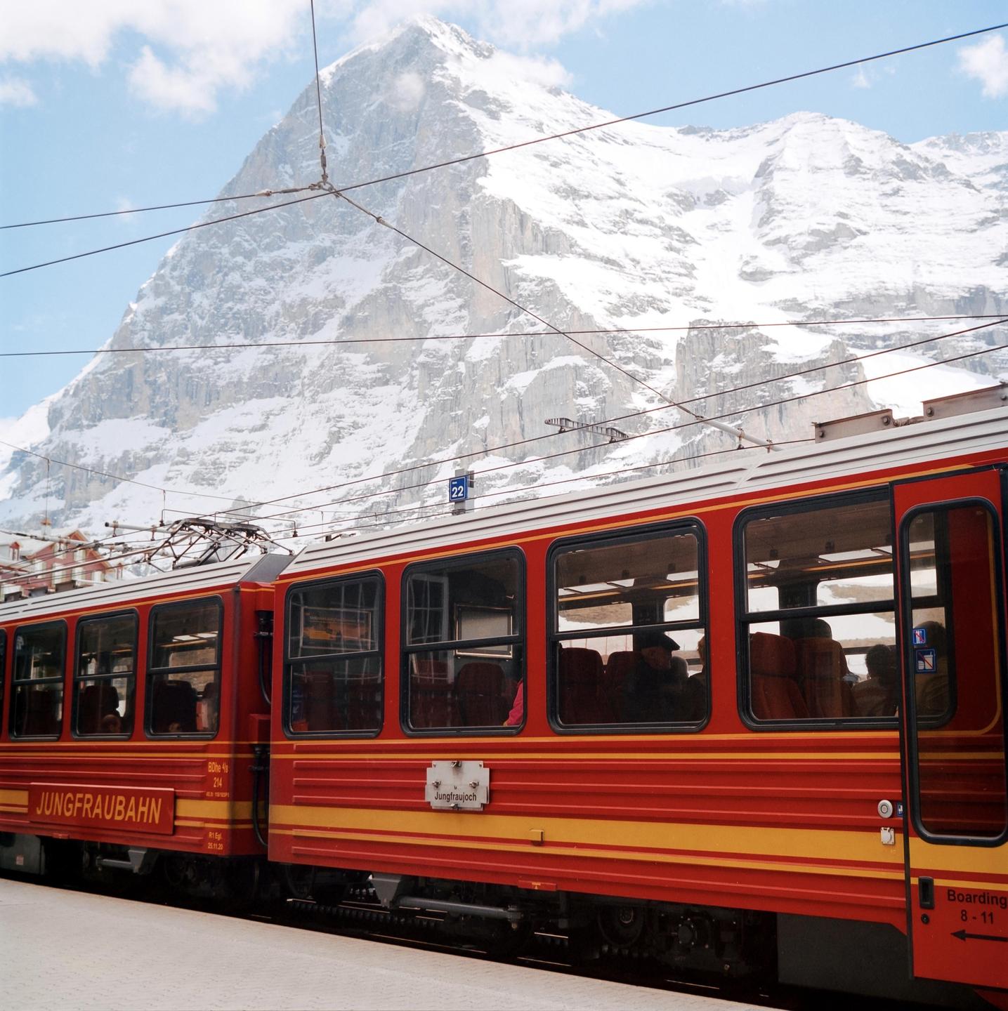 A red and yellow train is parked at a station with a snow-covered mountain in the background.