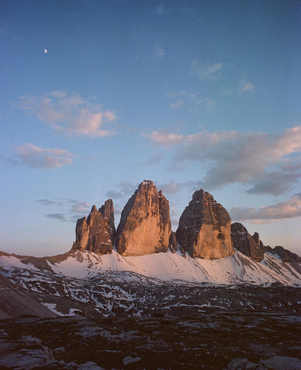 Three prominent mountain peaks are illuminated by the setting sun, with snow on their lower slopes and a crescent moon visible in the sky.