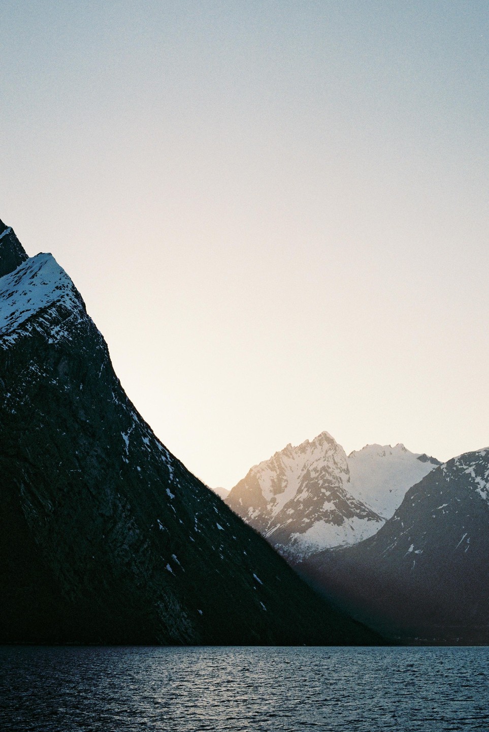 A body of water reflects the light from a snow-capped mountain range under a clear sky.