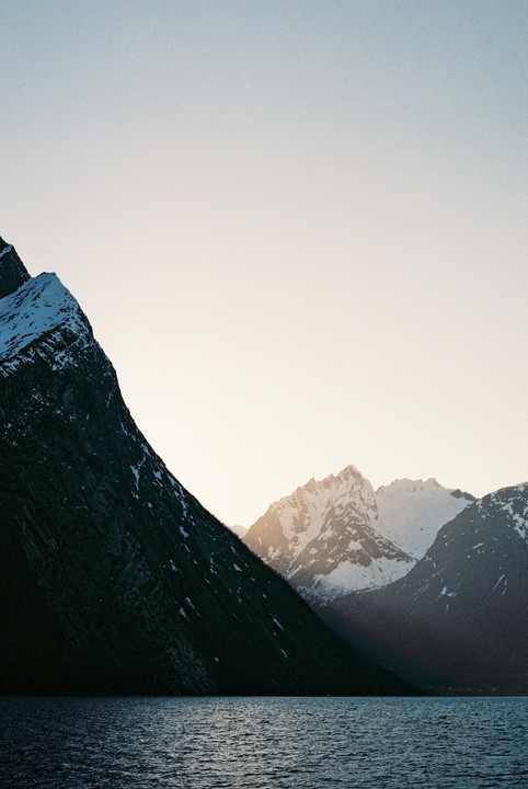 A body of water reflects the light from a snow-capped mountain range under a clear sky.