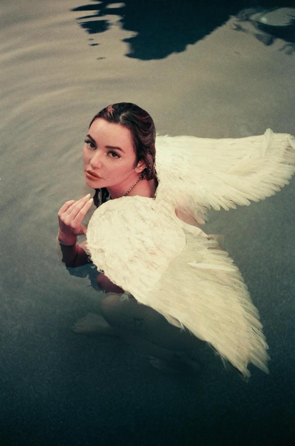 A woman with large white wings is partially submerged in water, looking towards the viewer.