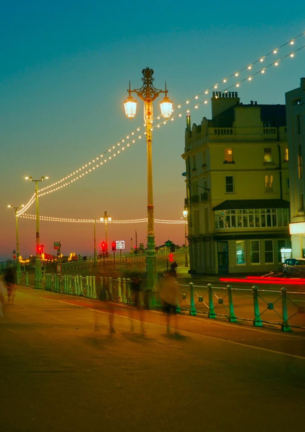 A street scene at dusk features a large ornate lamppost and strings of lights illuminating a building and blurred figures walking along a road.