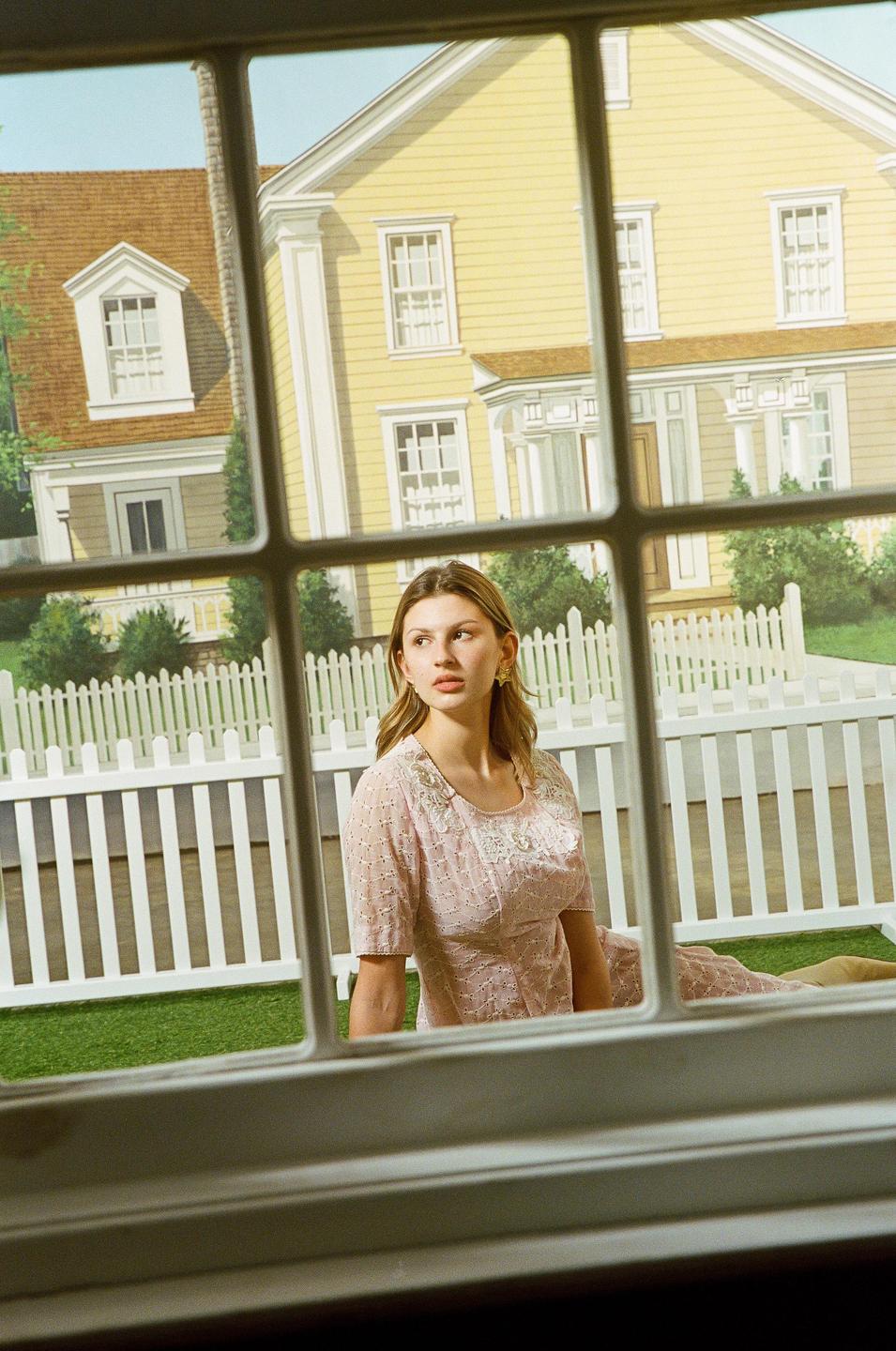 A woman in a pink dress sits in a staged suburban setting with a yellow house and white picket fence visible through a window.