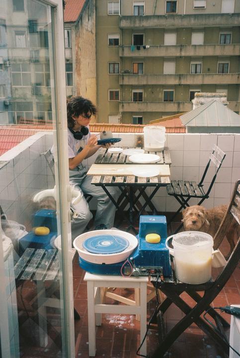A woman works on pottery at a table on a balcony with a dog nearby.