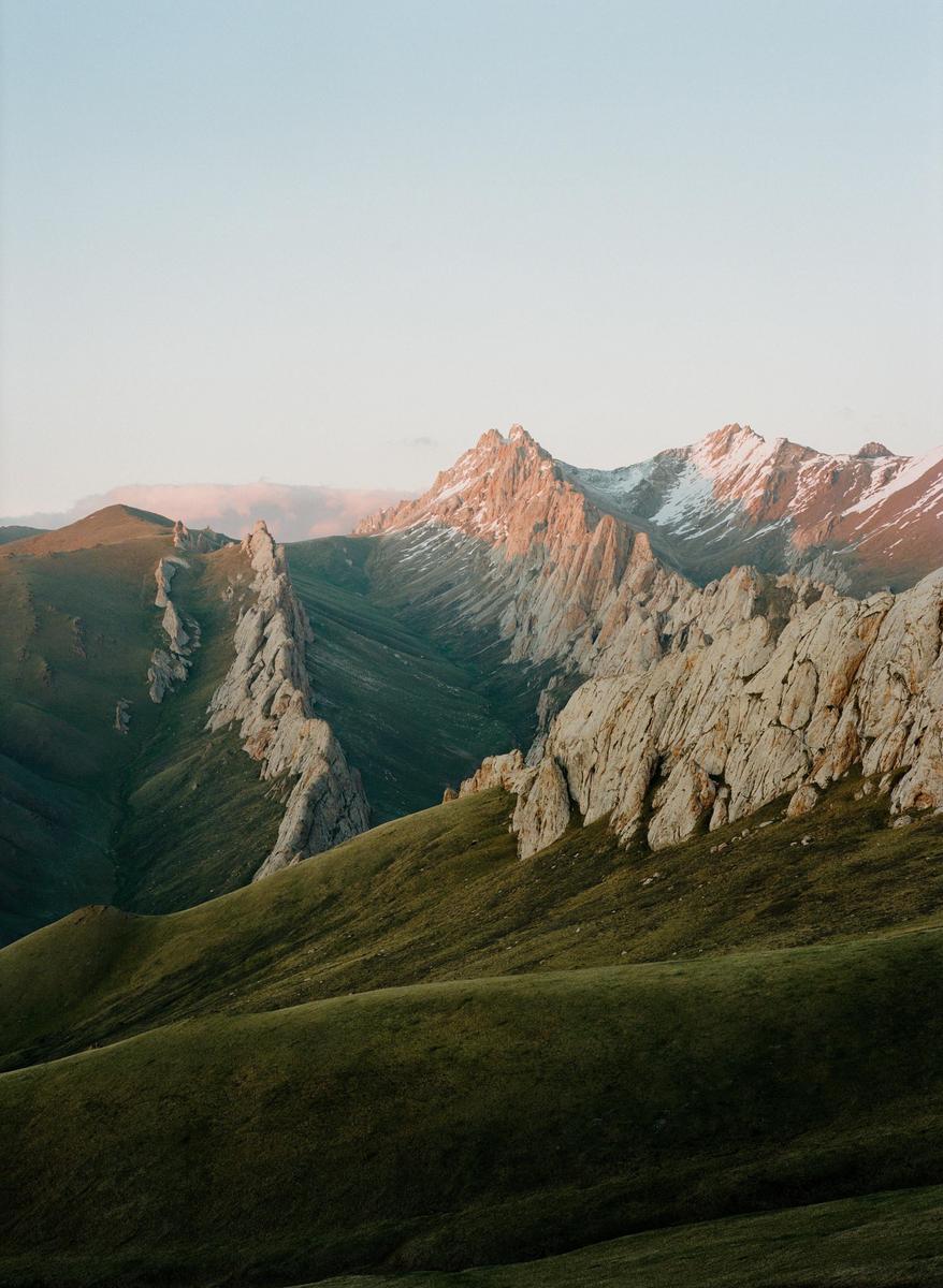 Sunlight illuminates the peaks of a mountain range with green hills in the foreground.