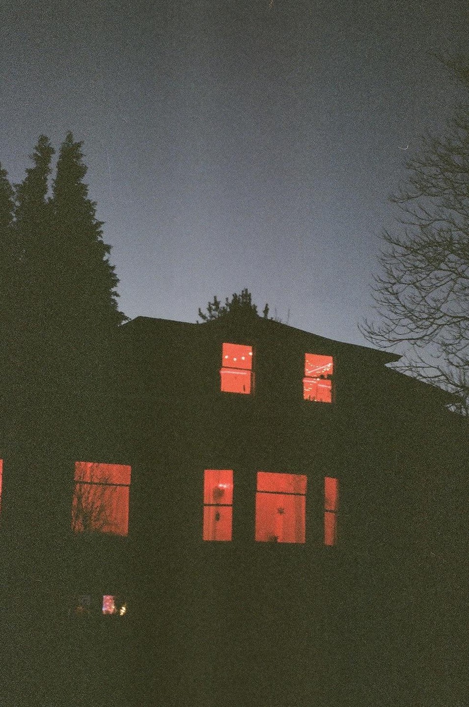 A dark house with red glowing windows stands against a dark sky with a sliver of moon.