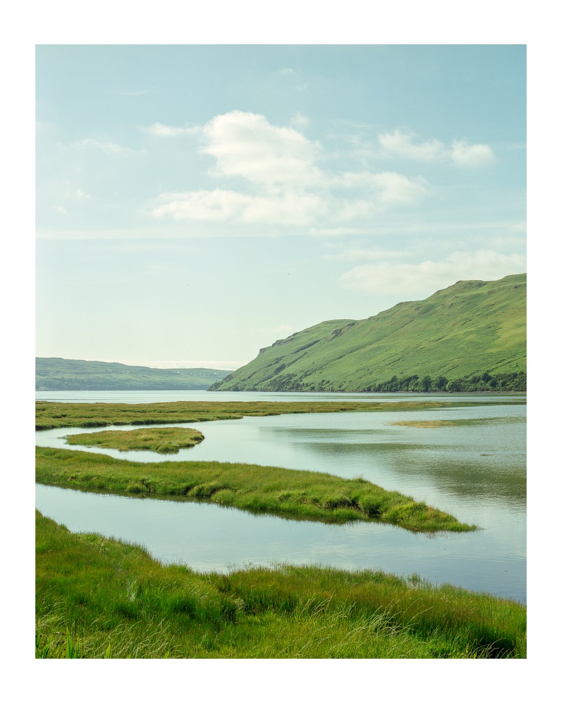 A body of water with winding grassy islands is surrounded by green hills under a light blue sky with scattered clouds.