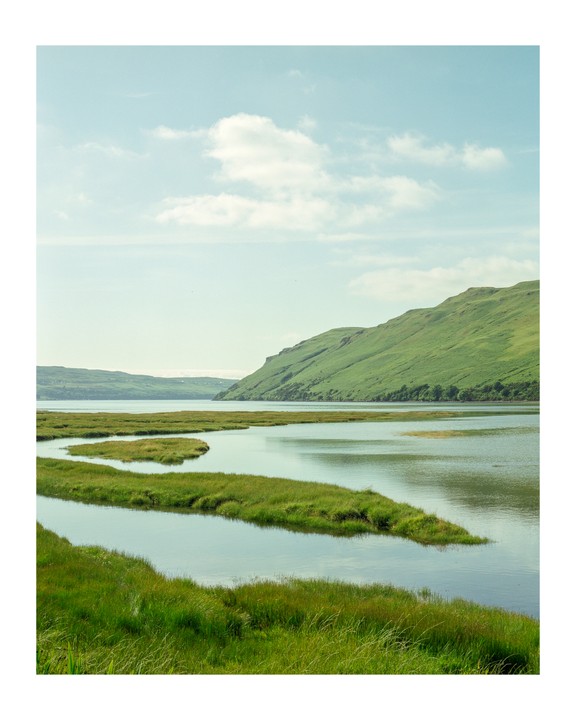 A body of water with winding grassy islands is surrounded by green hills under a light blue sky with scattered clouds.