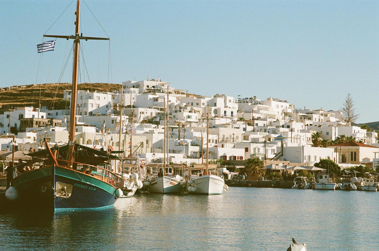 Boats are docked in a harbor with a town of white buildings on a hill in the background.