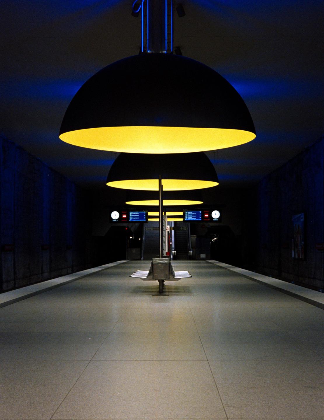 A subway platform is illuminated by a row of large, yellow, dome-shaped lights and blue ambient light.