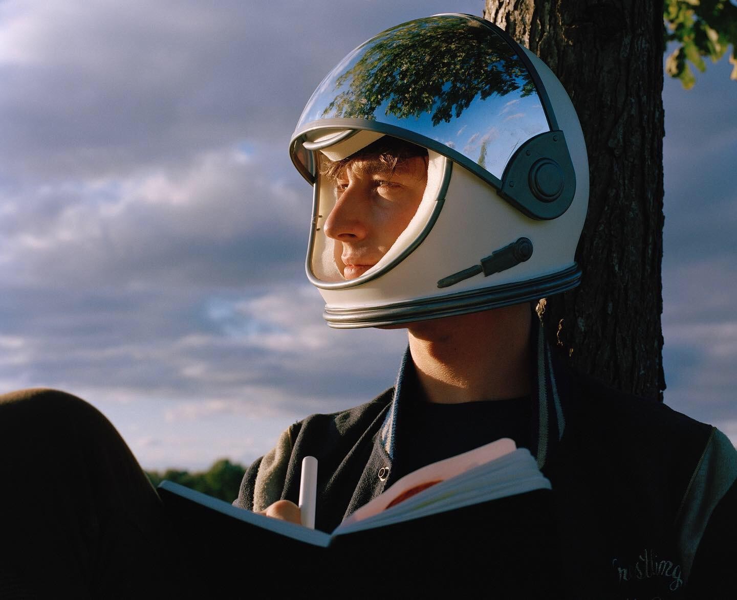 A man wearing a space helmet is writing in a notebook while leaning against a tree.