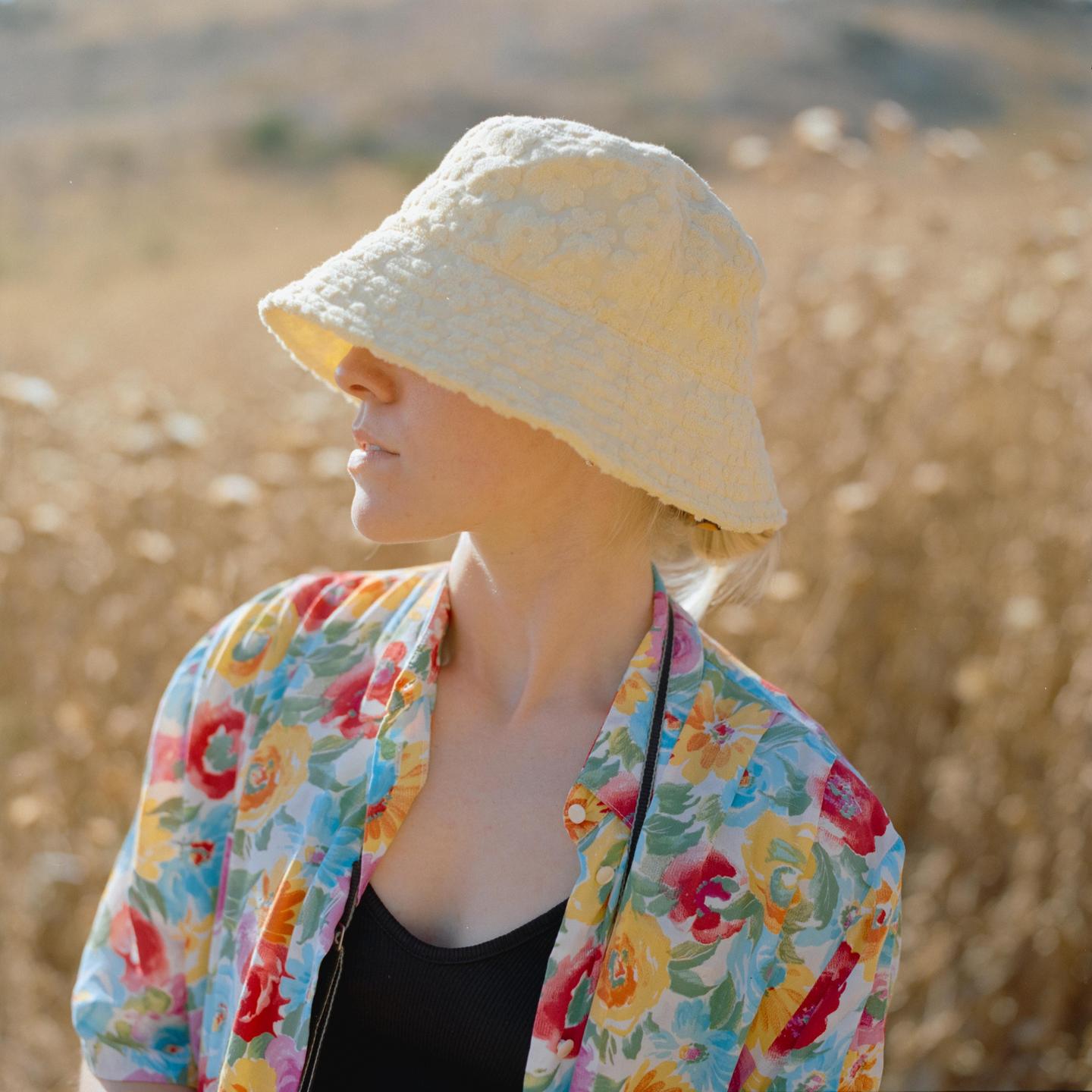 A woman in a floral shirt and bucket hat looks to the side in a field of dry grass.