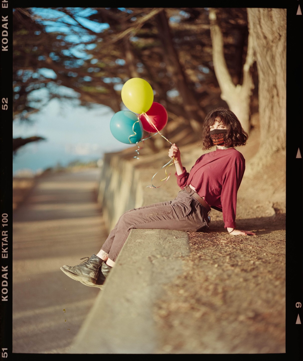 A woman sits on a concrete ledge holding three balloons.