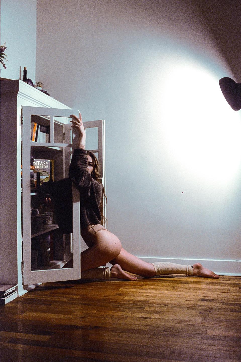 A woman in a sweater and underwear poses on a wooden floor next to a white bookshelf.