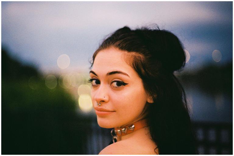 A woman with dark hair and a studded collar looks over her shoulder at the viewer.