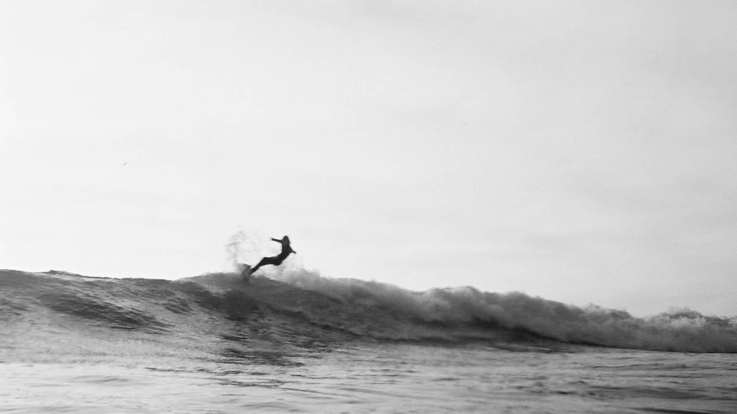 A surfer rides a wave, captured in a black and white, wide shot.