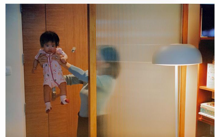 A baby in a pink onesie is held up by a woman whose reflection is visible in a fluted glass door.