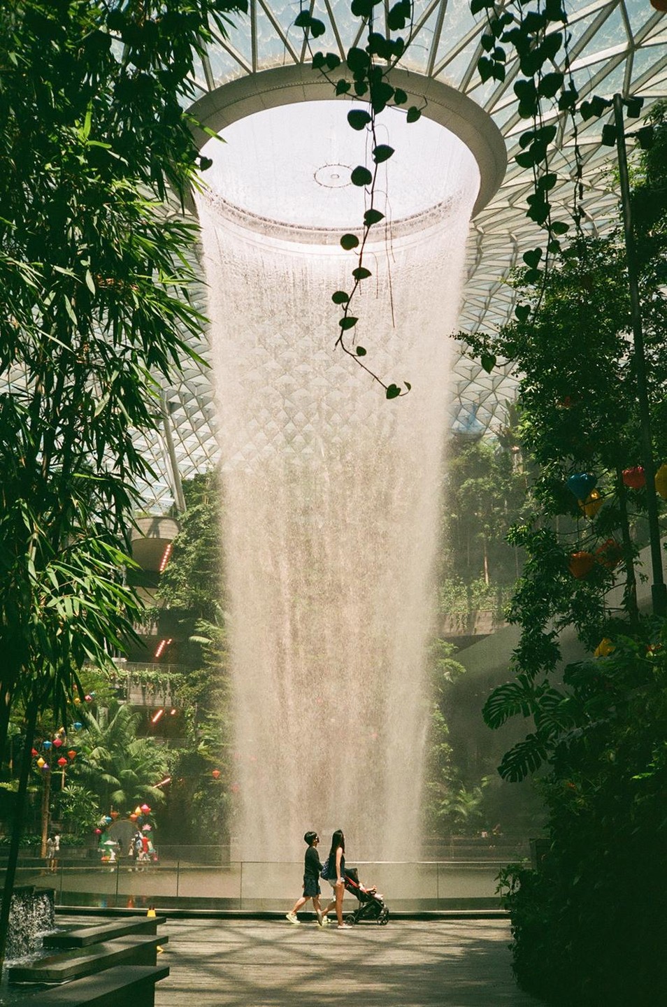 Two people walk past a large indoor waterfall surrounded by lush greenery and a glass dome.