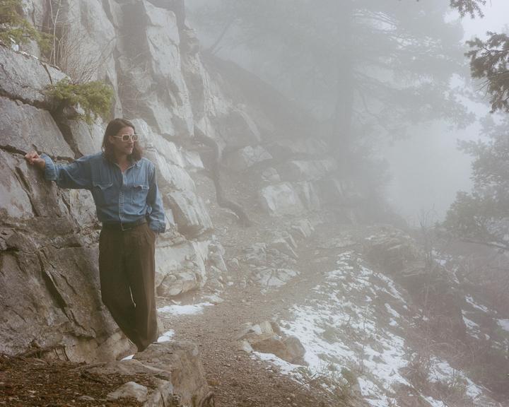 A man stands on a snowy mountain path next to a rock face in the fog.