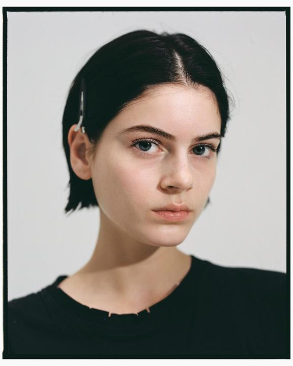 A woman with short dark hair and a black shirt looks directly at the viewer in a studio portrait.
