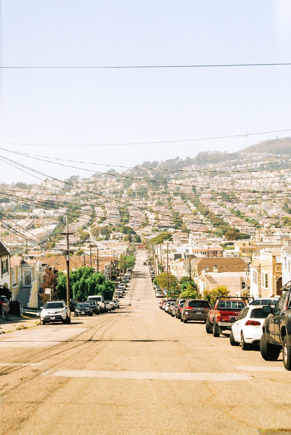 A long, steep street is lined with parked cars and houses, leading up to a hill covered in more houses.