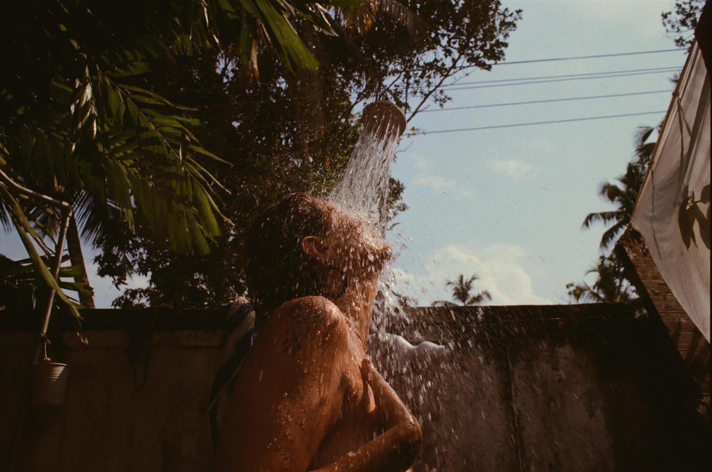 A person showers outdoors under a stream of water from a coconut shell, surrounded by lush foliage and a clear sky.