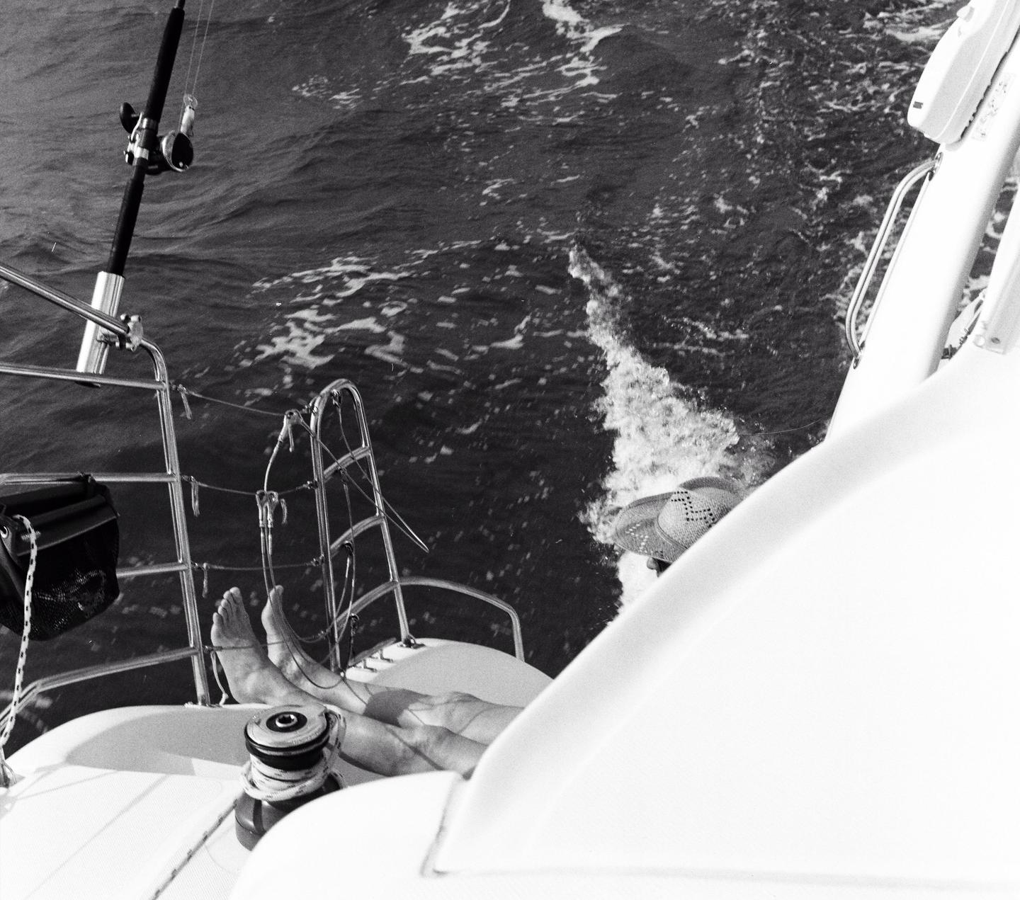 A person's legs and feet are visible on a boat deck, with a fishing rod and the ocean in the background.