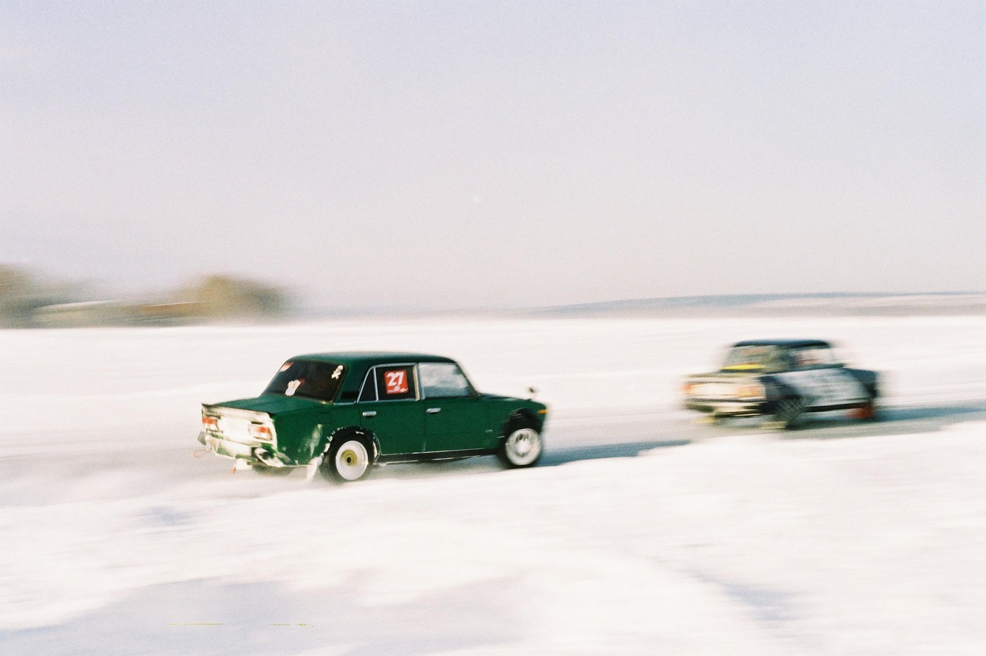 Two cars race across a snow-covered landscape, with the lead green car kicking up snow.