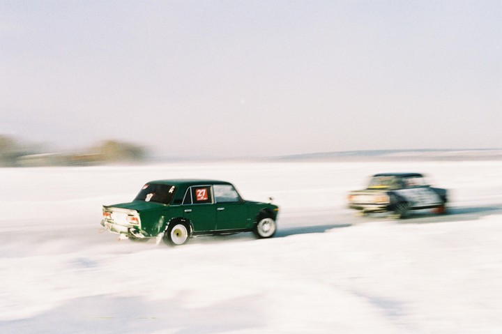 Two cars race across a snow-covered landscape, with the lead green car kicking up snow.