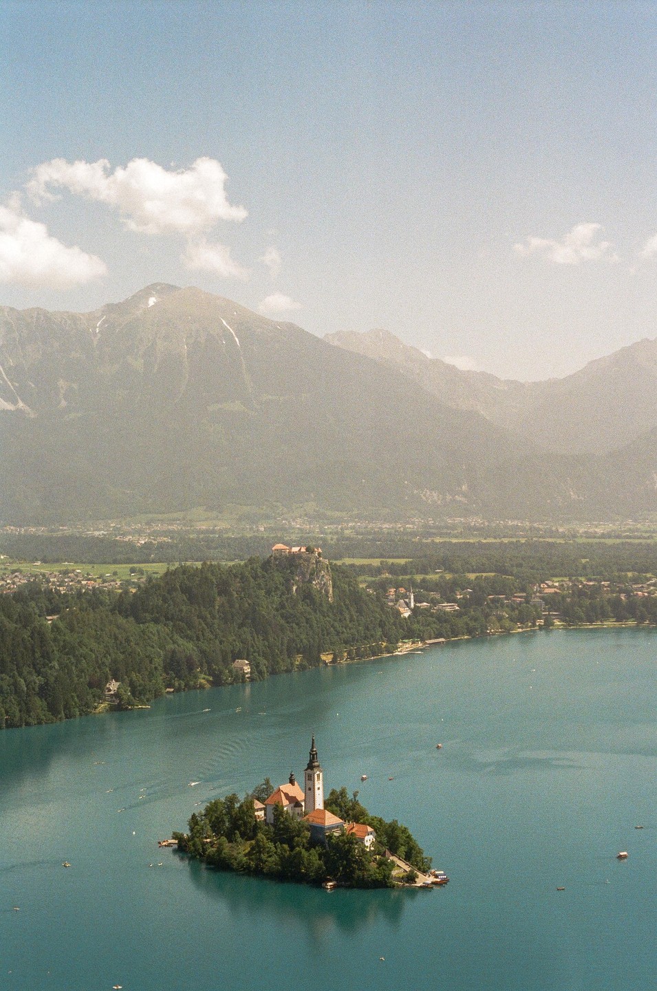 A small island with a church sits in a lake with mountains and a castle in the background.