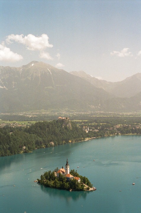 A small island with a church sits in a lake with mountains and a castle in the background.