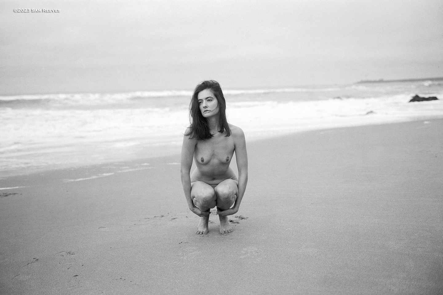 A woman crouches on a sandy beach with waves in the background.