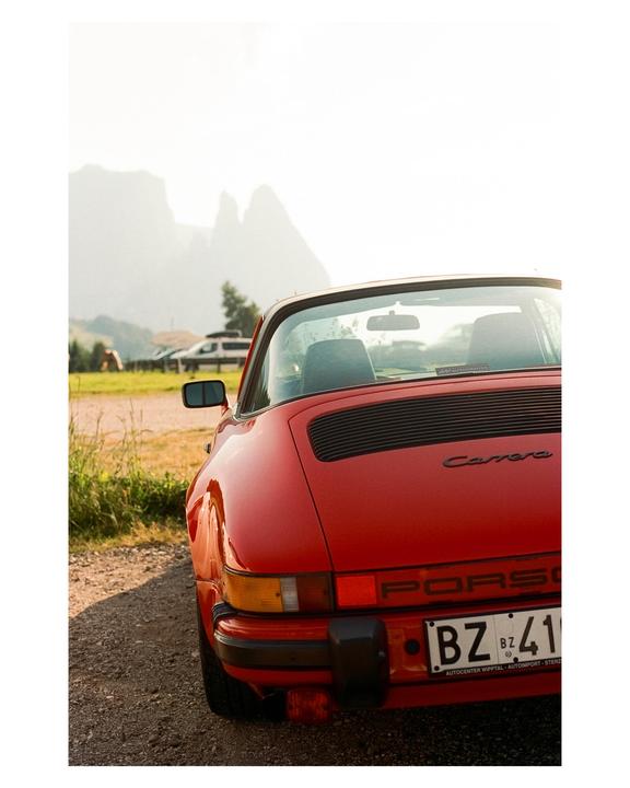 A red Porsche is parked on a gravel road with mountains in the background.