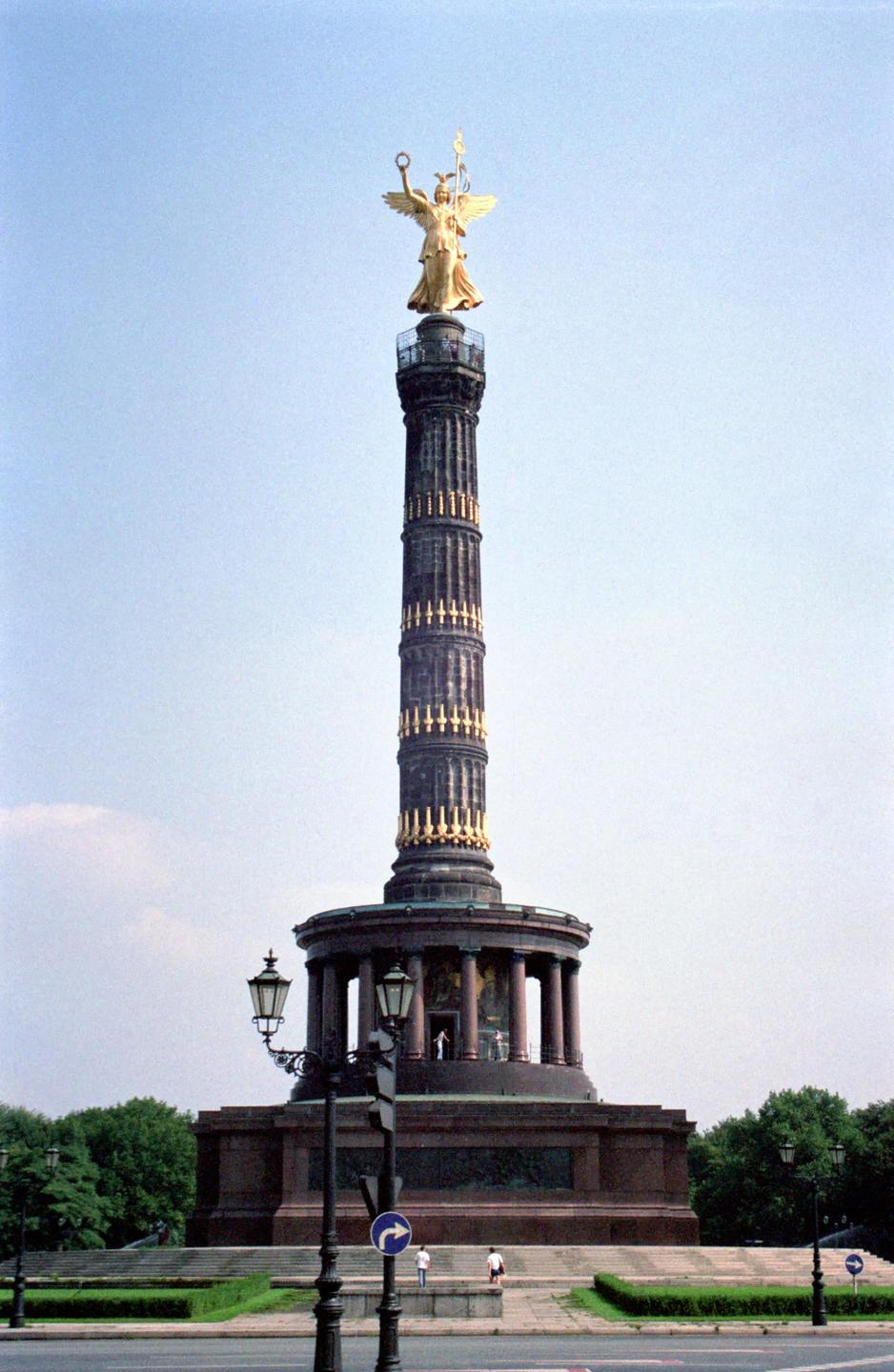 A golden statue of an angel stands atop a tall, dark column with a circular base, surrounded by trees and streetlights.